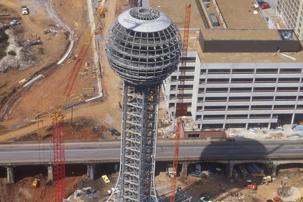 A high-angle historical photograph showing the Sunsphere under construction in Knoxville, Tennessee, for the 1982 World's Fair. The structural steel framework of the tower and the unfinished sphere are visible, surrounded by construction cranes, dirt lots, and neighboring buildings in progress.