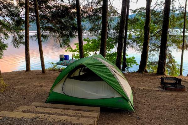 A green and white tent sits beside a lake with tall trees and sunrise colors reflecting off the water.