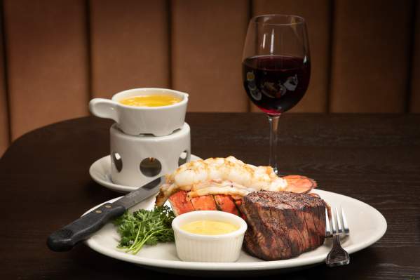 A delicious steak and lobster tail on a plate with a glass of wine to the side.
