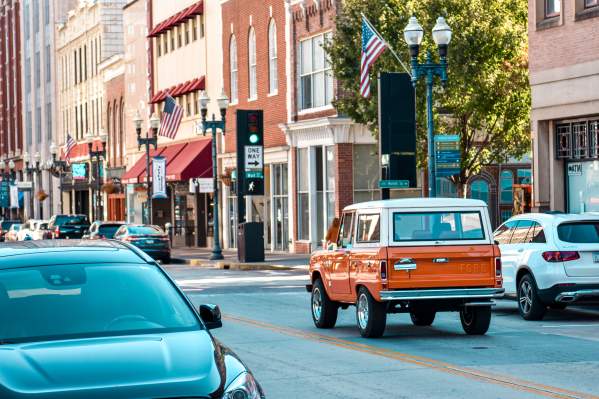 A street-level view of historic Gay Street in downtown Knoxville, Tennessee, featuring a classic orange and white Ford Bronco driving away from the camera. The urban streetscape is lined with brick buildings, American flags, and streetlights, with other modern vehicles in the foreground and background under soft, daytime lighting.