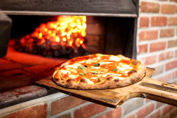 A perfectly cooked pizza is being pulled out of a woodfire oven on a wooden paddle.