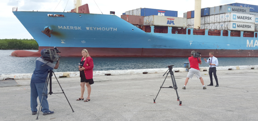 Photo of two reporters and videographers reporting from dockside with large cargo ship in the background.
