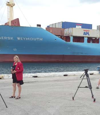 Photo of two reporters and videographers reporting from dockside with large cargo ship in the background.