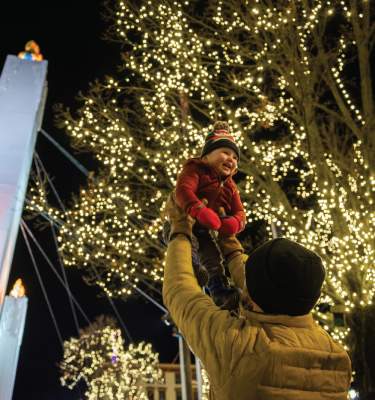 A smiling child hoisted in the air among Christmas lights and the Peace Candle at Easton Winter Village