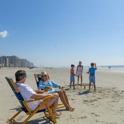 A mom and dad relax on the beach while their children play a beach game in the background.