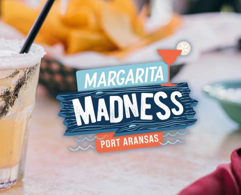 A margarita sitting on a table with chips int he background. To the right of the margarita is a blue and orange logo that says Margarita Madness Port Aransas