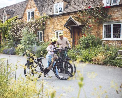 A couple have dismounted their bikes in front of a stunning cottage, with red roses climbing over it's porch, and are looking at their map