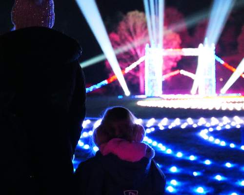 A mother and small child stand with their backs to the camera looking at a small, lit up London Bridge. There are roaming spotlights & glowing fairylights on the floor in all different colours. The trees off in the distance are also lit up in blue and pink.