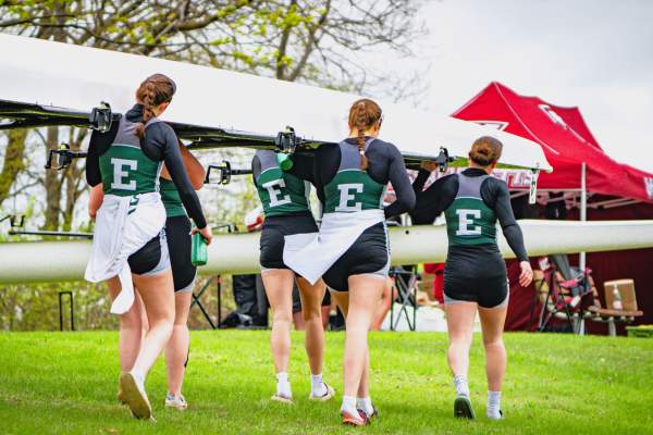 Team of five Eastern Michigan University Womens rowing teammates carrying large rowing boat as they walk across grassy area.