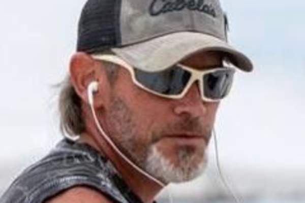 Headshot of a man in wraparound sunglasses, a tank top, a baseball cap, and with wired headphones in looks over his shoulder at the camera.