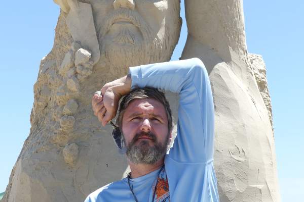 A man in a light blue long sleeve shirt stares off camera with his hand over the top of his head and holding a spade against his face. He is mirrored by the larger-than-life sand sculpture directly behind him.
