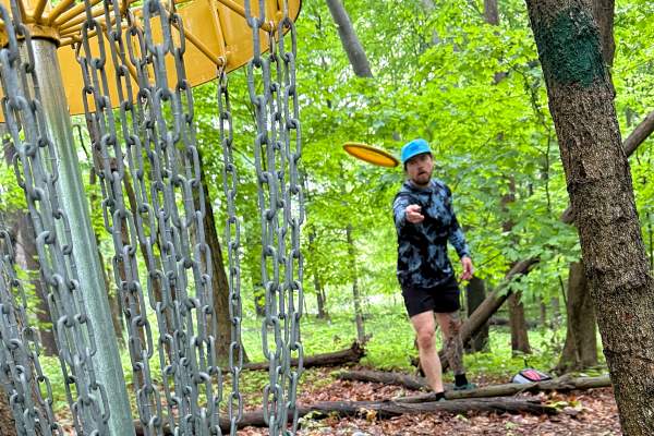 Man throws disc golf frisbee towards camea, which sits behind the hole in heavily wooded and shaded area