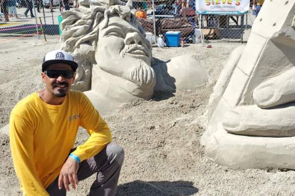 A man in a yellow long sleeve shirt kneels down next to a sand sculpture depicting a man's face and hand coming up out of the ground, rocking out on a guitar