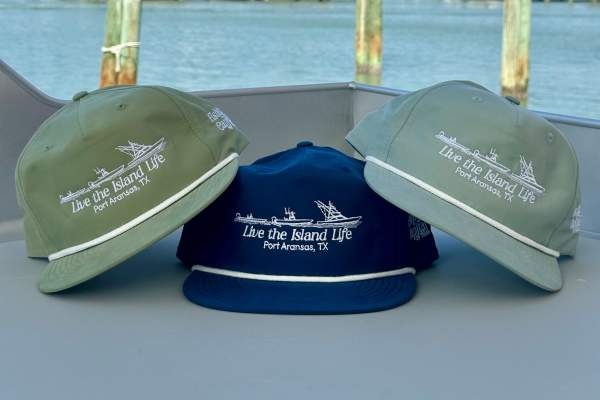 Three "Fishing Capital of Texas" hats in green and blue sitting on a table near a sunny harbor.