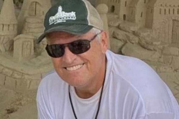 A middle-aged man in a baseball cap, a long-sleeve white shirt, and dark sunglasses leans over in front of a sand sculpture and smiles at the camera.
