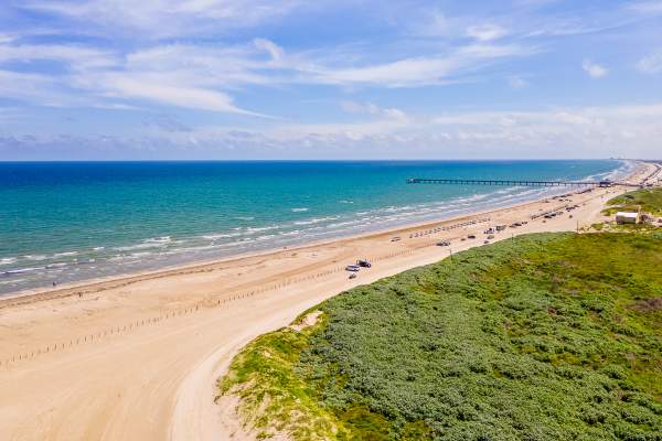 Drone image of a beach that shows the water, beach, and sand dunes from very high. Down the beach, a few cars and a pier are visible.