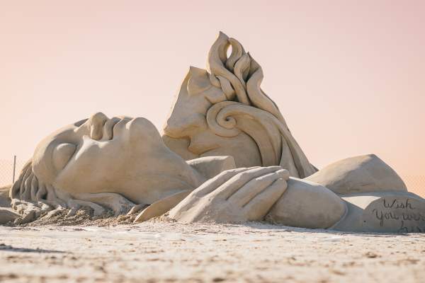 Sand sculpture depicting a woman lying on her back with her hand over her breasts. Half the face of a man is visible kissing her neck