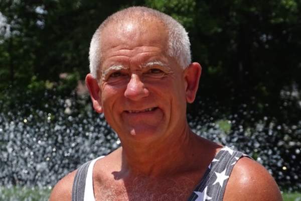 A tanned older man with white hair smiles at the camera. He's outside in front of some greenery and he's wearing a stars and stripes tank top