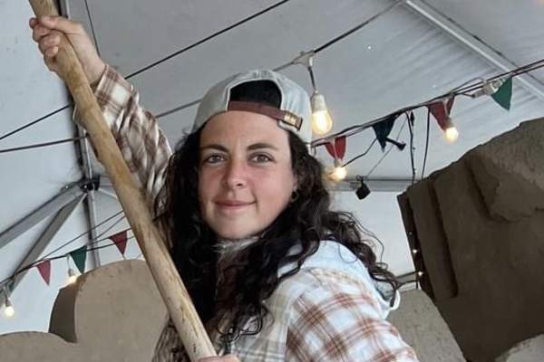 A woman wearing a baseball cap and a flannel shirt holds up a shovel like a jousting weapon, smiling at the camera in front of a half-carved pile of sand.