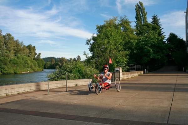 A person on a three-wheeled recumbent bike rolls down the wide smooth paved path alongside the Willamette River. The cyclist is wearing warm weather clothing. The sky is blue and the trees are bright green.
