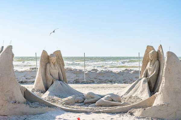 Sand sculpture depicting four angels at four corners holding a blanket. In the middle of the blanket, a woman lies curled up.