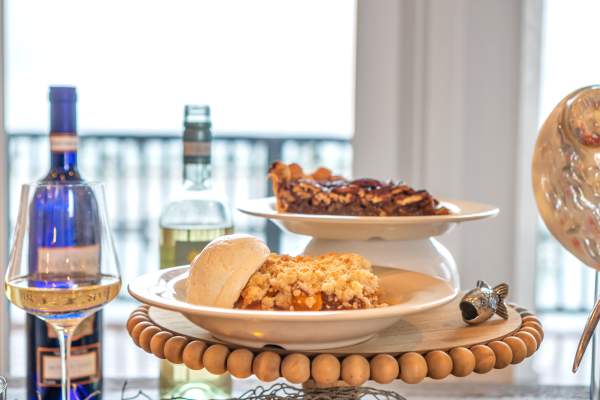 A plate of cobbler with ice cream and a plate of pecan pie sit stacked on an elevated dessert tray. To the left are open bottles and glasses of wine.
