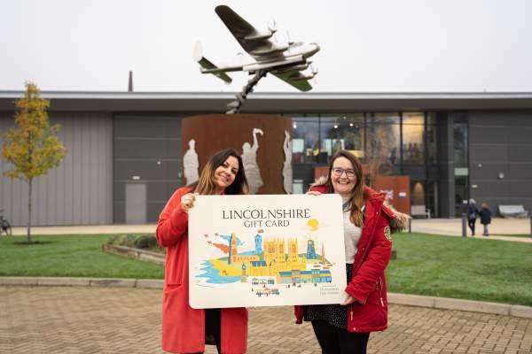 Two people holding a large Lincolnshire Gift Card in front of the International Bomber Command Centre