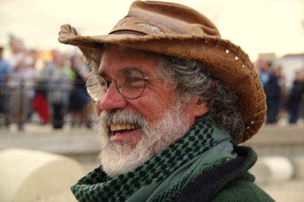 Portrait of an older man with a beard and glasses smiling off to the left of the camera. H e is wearing a battered floppy hat and a green neck scarf.