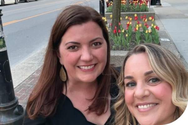 two women smile for the camera. They stand outside near a street with red and yellow tulips blossoming in the background, in the sidewalk planters.
