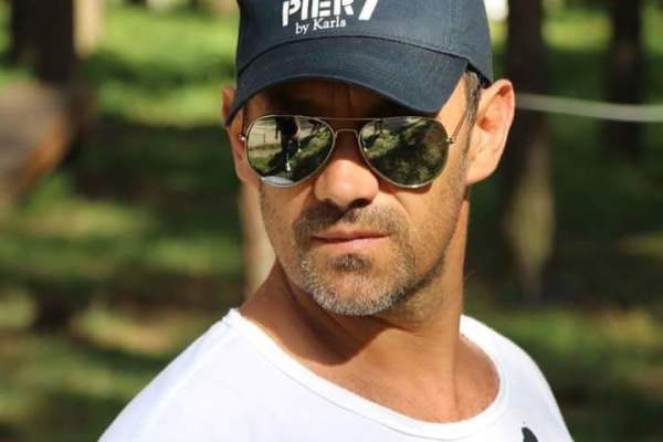 Chest up photo of a man wearing a white T-shirt with a black headphone design. He also has on mirrored sunglasses and a baseball cap.