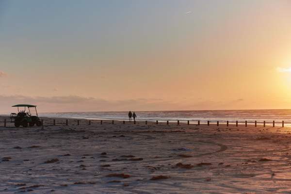 A silhouetted couple walks along the beach at sunrise. A golf cart is parked in the distance.