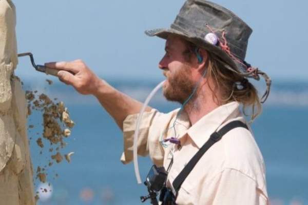 A man in a hat and a ponytail holds a carving tool in one hand to his sand sculpture and has a blowing straw in his mouth.