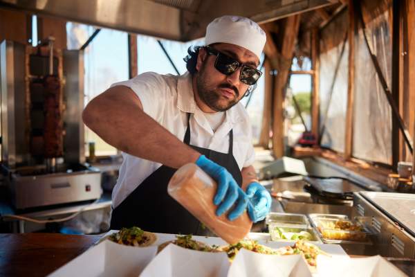 A man in an apron and chef's hat squeezes sauce from a bottle over a row of tacos