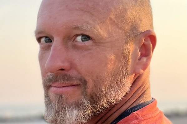 A bald, bearded man with blue eyes looks over his shoulder and smiles at the camera. This is a close up of his shoulders up. he's wearing a coral-colored shirt