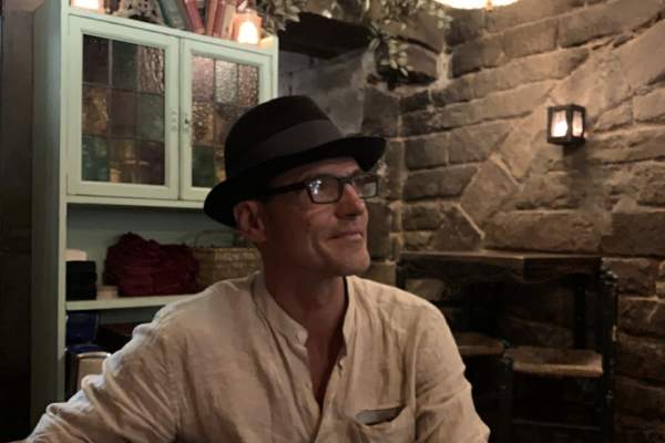 A man sits at a table in what looks like a restaurant smiling off to the right of the camera. He's wearing a hat and a buttoned linen shirt.