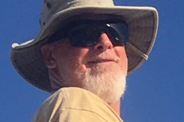 Photograph of a man with a white beard, sunglasses, a yellow T-shirt, and a wide-brim hat looks over his shoulder to the camera.