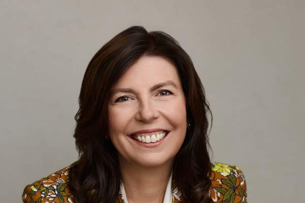 Headshot of Leslie Bruce. She's sitting, smiling at the camera with her hands crossed. She's wearing a white blouse and a floral-print blazer.