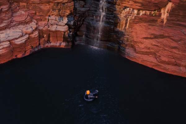 A person in an inflatable tube and canyoning gear floating at the bottom of a tall waterfall in Karijini National Park.
