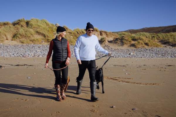 A man and woman wearing warm clothes, each walking a dog on a beach.