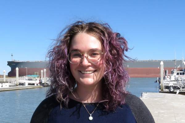 Woman with purple curly hair stands in front of a ship channel and smiles at the camera