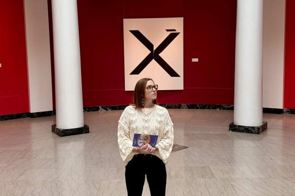 Woman looks away from camera at paintings that surround her that hang on walls.
