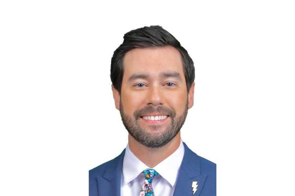 Headshot of a man in a suit with a dark beard and hair smiling at the camera. He wears a fun floral tie and a lightning bolt pin.