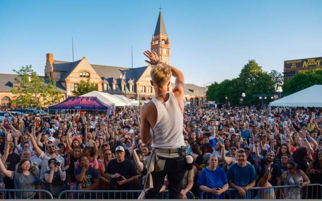A musician performs to a large, enthusiastic audience gathered at Cheyenne Depot Plaza. The crowd stretches across the plaza, with many people raising their hands and phones. The Cheyenne Depot clock tower stands prominently behind the audience on a bright, clear day.