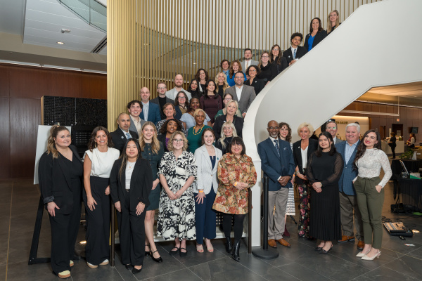Large group of professionals posing on a curved staircase inside a modern building with wood slat walls and glass railings.