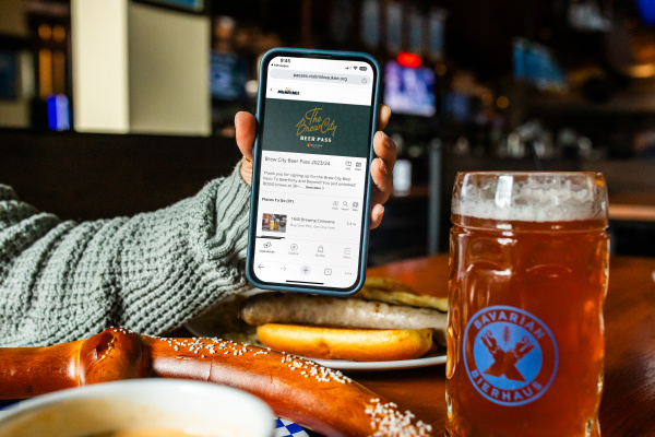 a hand holding up a phone displaying the Brew City Beer Pass over a table with a Bavarian Pretzel and a large pint of beer