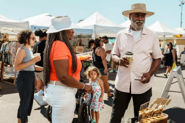 Visitors browse vendor booths at an outdoor market on a sunny day. Two people chat while holding coffee near a handcrafted goods display, as families and shoppers explore the bustling festival in the background.