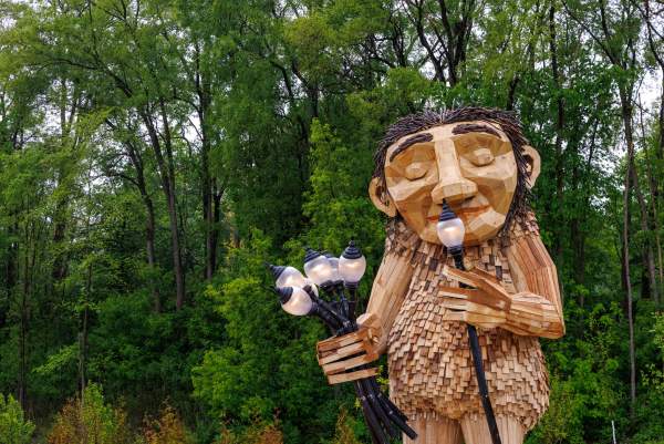 Large Wooden Troll sculpture smaelling lamps as if they were flowers against green trees.