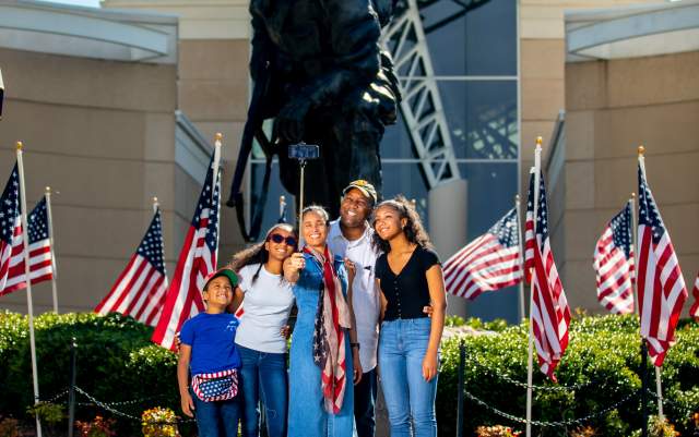 Family posing together in front of a large military statue with American flags displayed outside a museum building