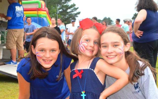 Three girls smiling together at the Stedman Fireworks Festival, with face paint and festival activities in the background.
