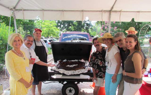 Group of adults smiling and gathered under a tent around a barbecue grill with smoked meat during an outdoor reunion-style event, creating a casual and social atmosphere.